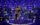The six queens of SIX: The Musical take center stage, each dressed in dazzling, futuristic Tudor-inspired costumes. They sing into handheld microphones with energetic expressions and dynamic poses, bathed in vibrant purple and gold stage lighting.