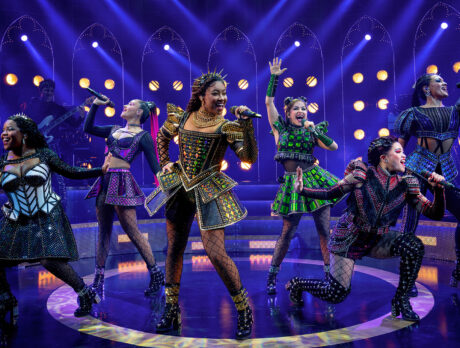 The six queens of SIX: The Musical take center stage, each dressed in dazzling, futuristic Tudor-inspired costumes. They sing into handheld microphones with energetic expressions and dynamic poses, bathed in vibrant purple and gold stage lighting.