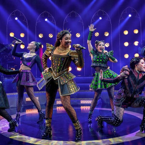 The six queens of SIX: The Musical take center stage, each dressed in dazzling, futuristic Tudor-inspired costumes. They sing into handheld microphones with energetic expressions and dynamic poses, bathed in vibrant purple and gold stage lighting.