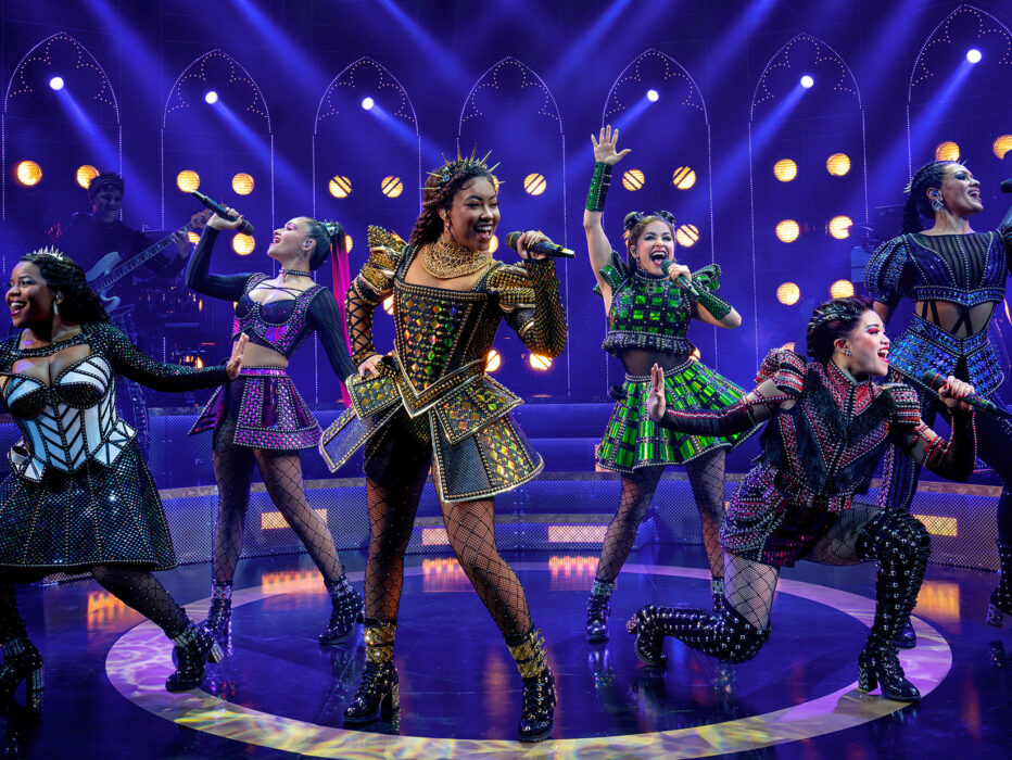 The six queens of SIX: The Musical take center stage, each dressed in dazzling, futuristic Tudor-inspired costumes. They sing into handheld microphones with energetic expressions and dynamic poses, bathed in vibrant purple and gold stage lighting.