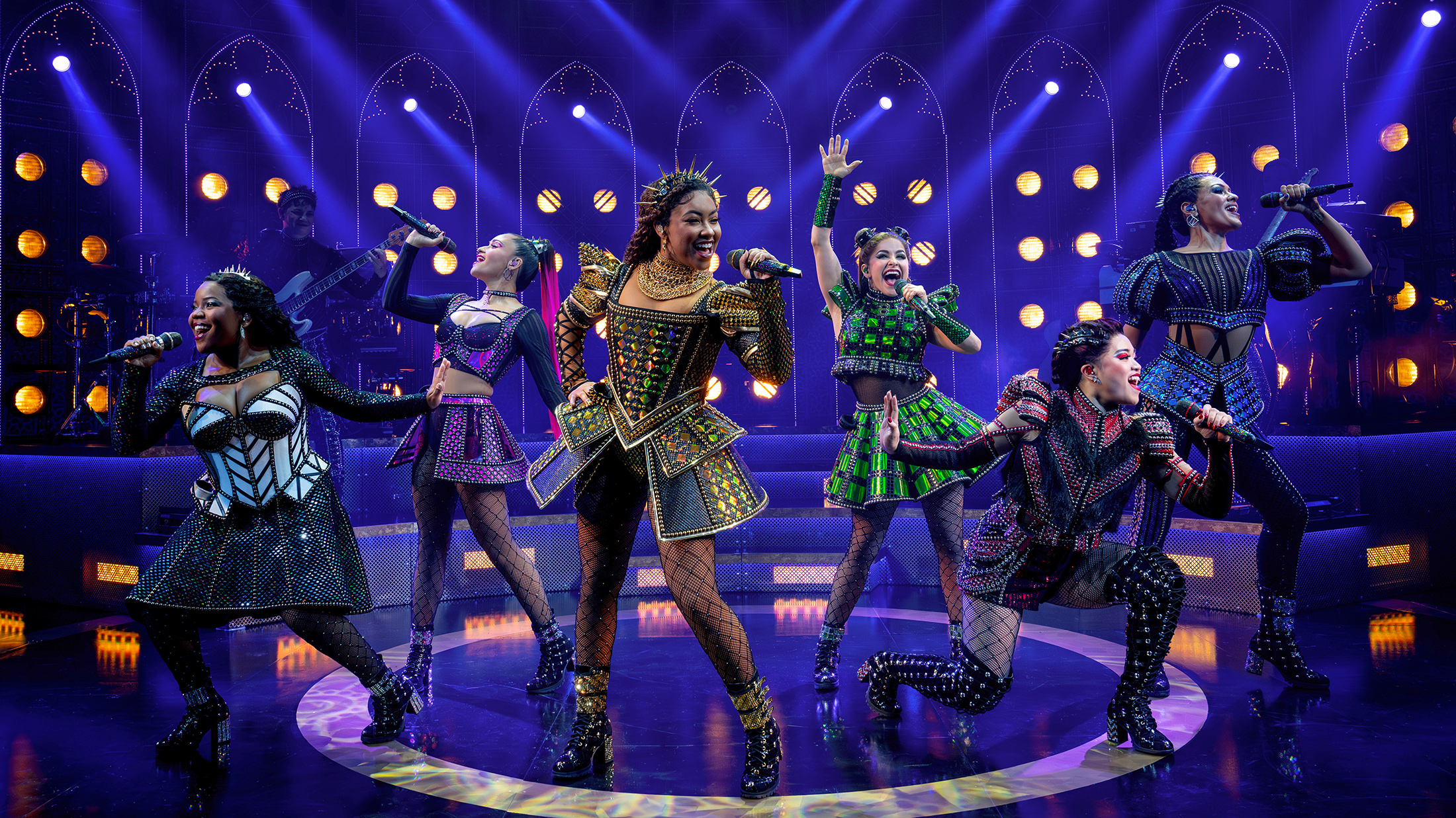 The six queens of SIX: The Musical take center stage, each dressed in dazzling, futuristic Tudor-inspired costumes. They sing into handheld microphones with energetic expressions and dynamic poses, bathed in vibrant purple and gold stage lighting.