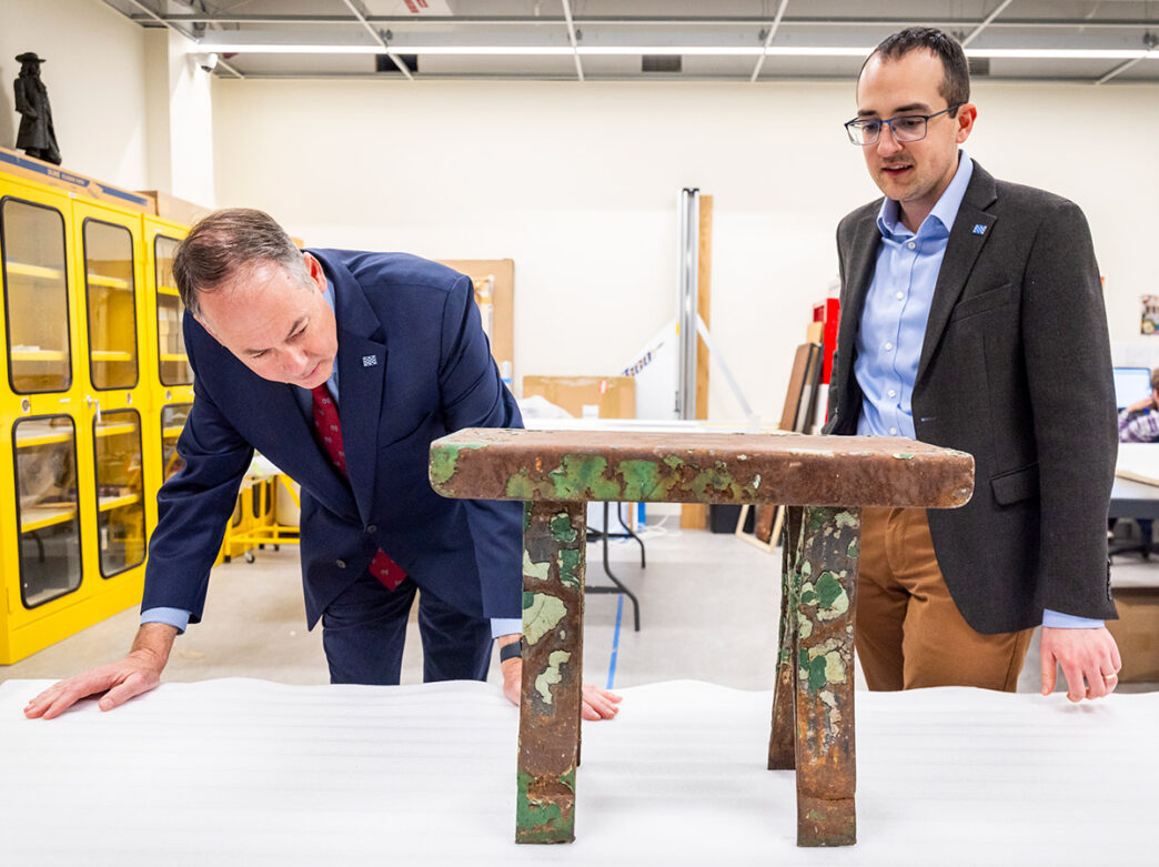 Two men in suits look at an old, metal prison bench used as part of a museum exhibition