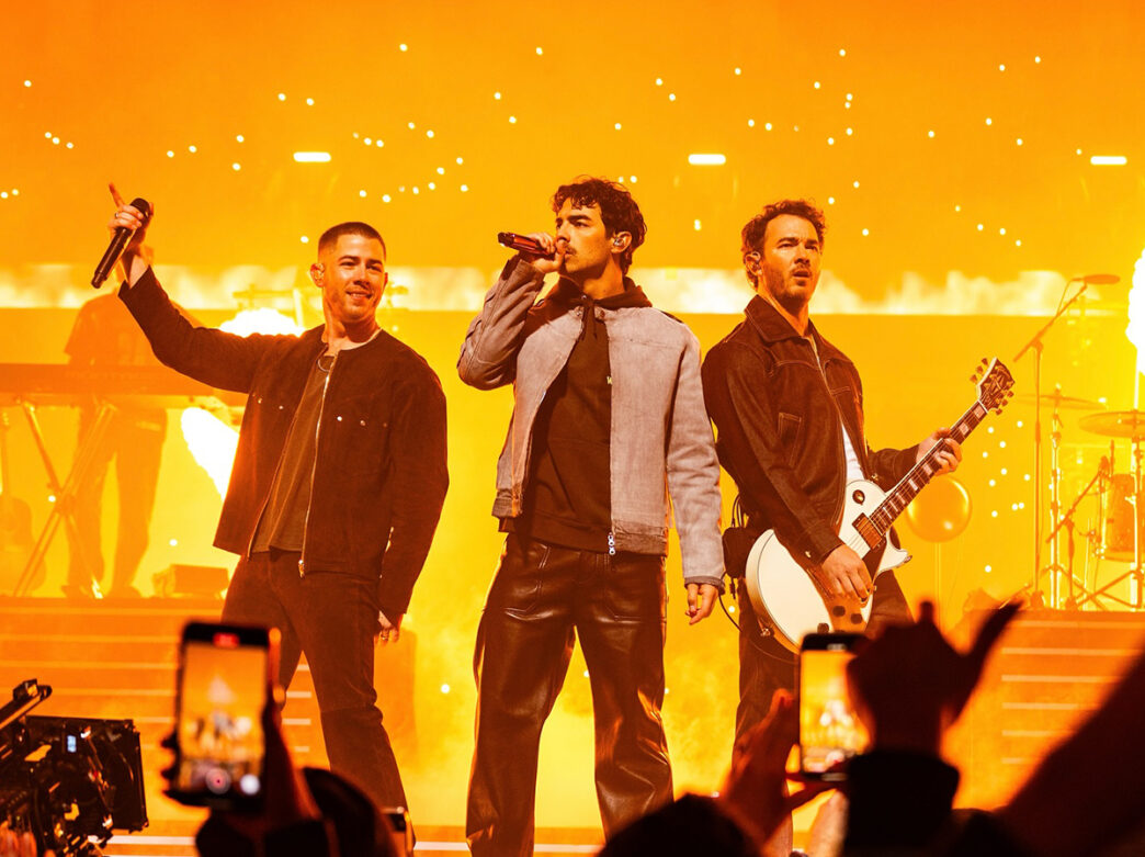 The three Jonas Brothers sing and play electric guitar on a brightly lit stage in front of a cheering crowd.