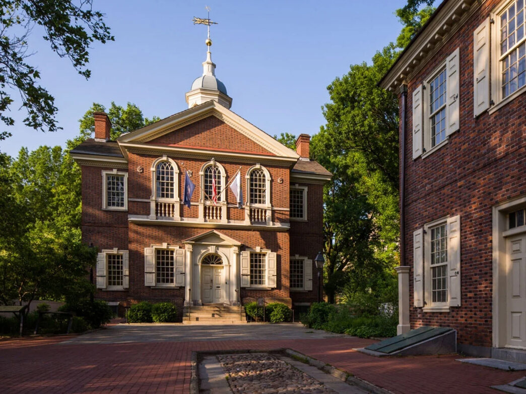 A historic red-brick building with white shutters, flags above the entrance and a small weather vane on top.