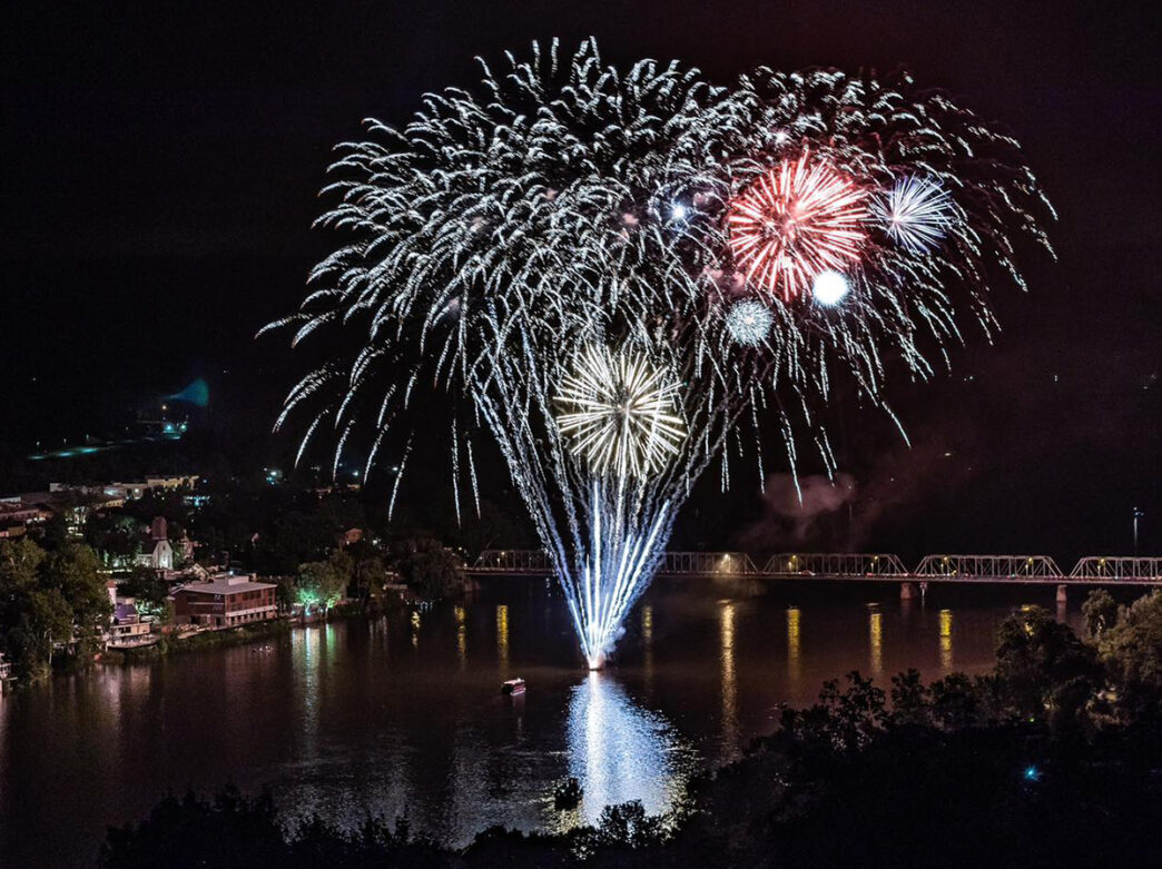 Colorful fireworks burst over the Delaware River in New Hope, reflecting off the water and lighting up the dark sky.