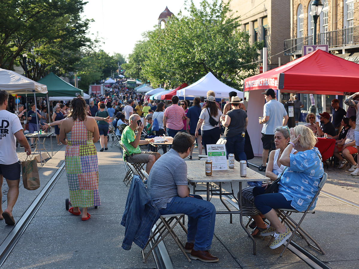 People walk, shop and dine at an outdoor street festival lined with colorful vendor tents and historic brick buildings on a sunny day.