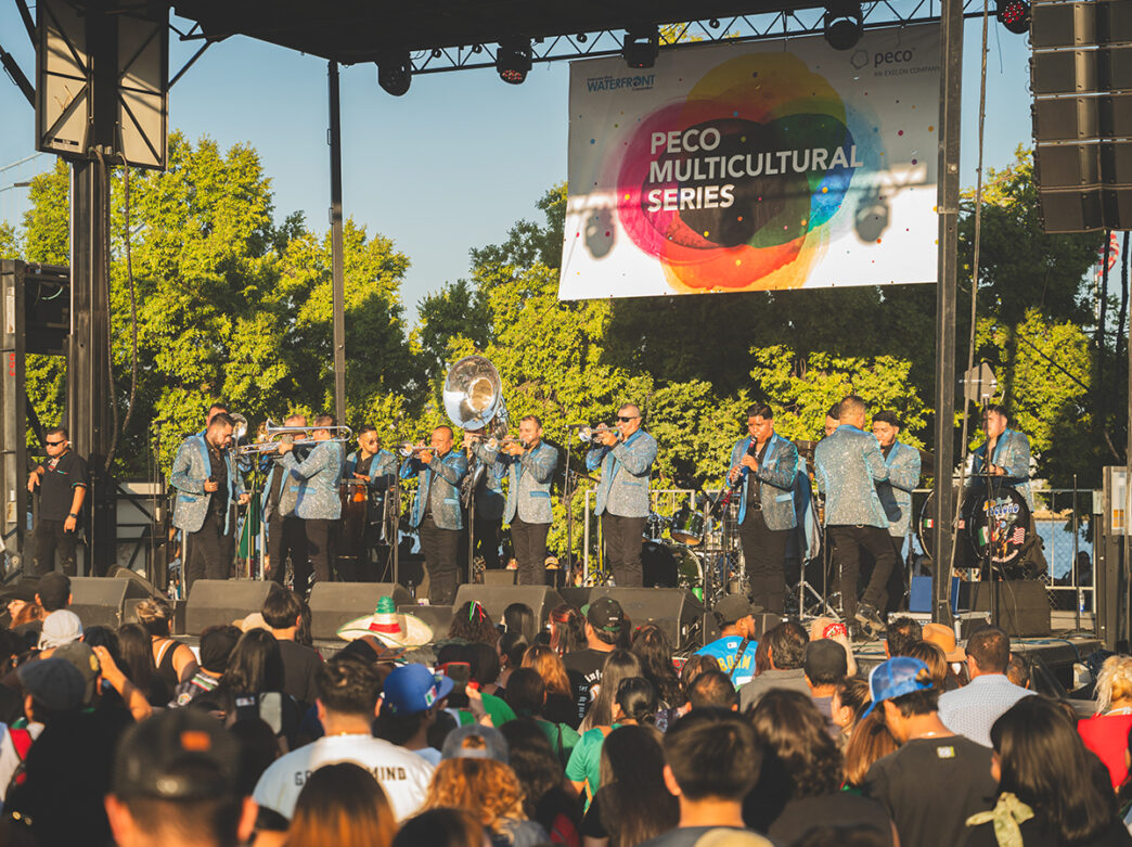 A mariachi band in matching blue jackets performs on stage before a large crowd at the Mexican Independence Day celebration.