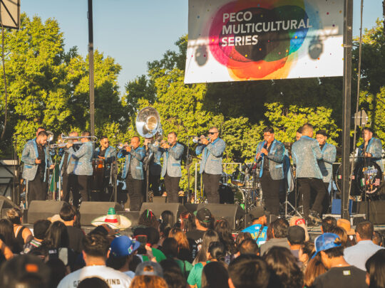 A mariachi band in matching blue jackets performs on stage before a large crowd at the Mexican Independence Day celebration.