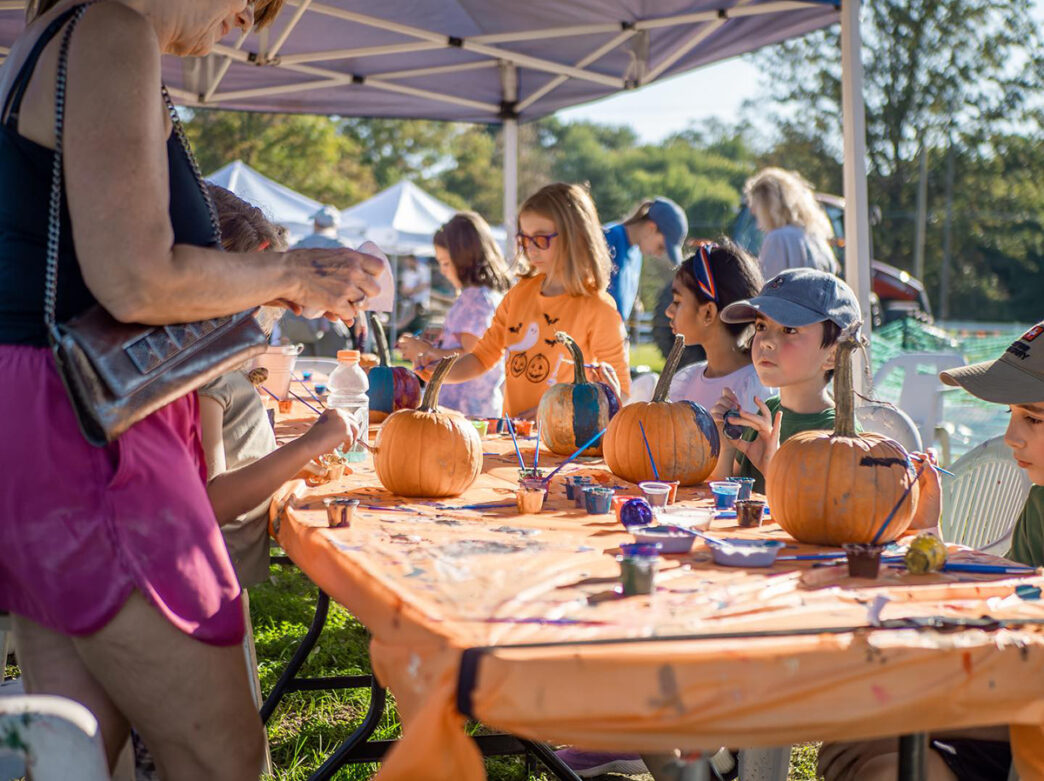 Children paint pumpkins at an outdoor tables underneath a pop-up tent during a fall harvest festival, surrounded by art supplies.