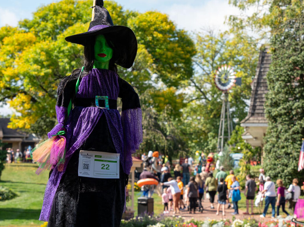 A witch themed scarecrow with a green face and purple dress stands on display at Peddler's Village. Visitors walking along brick pathways can be seen in the background.