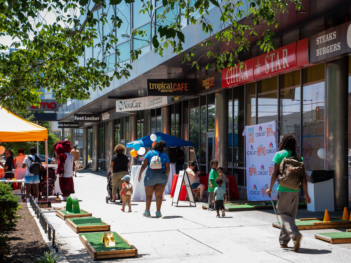 A group of people walking past storefronts on a bright summer day, mini golf, games and mascots are out