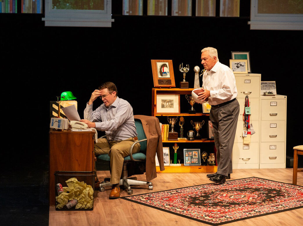 Two actors perform on stage in an office set, one sitting at a desk anxiously reading a piece of paper, while the other holds a football.
