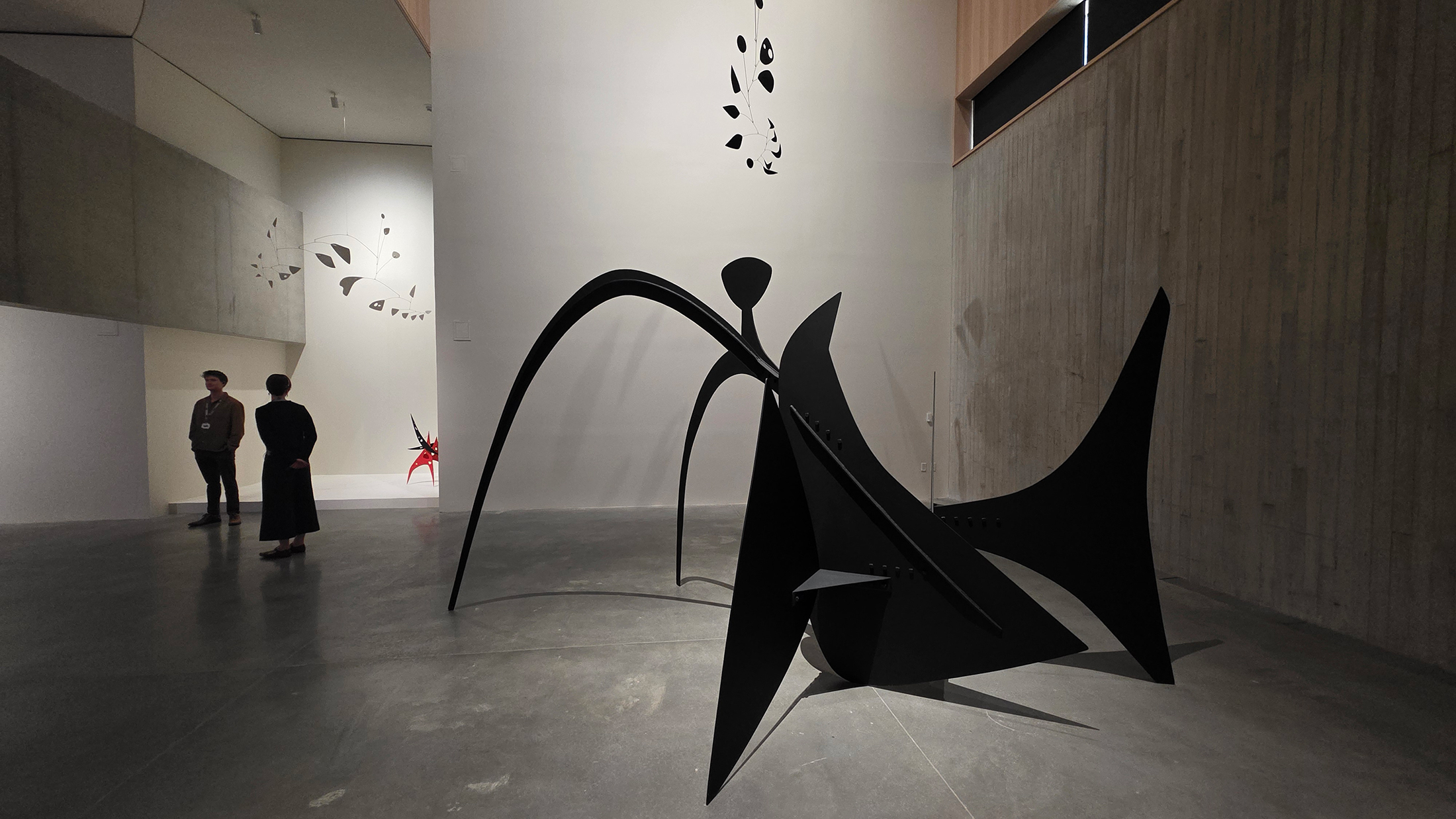 A large black abstract sculpture with sweeping, curved forms stands in a spacious gallery with visitors observing nearby.