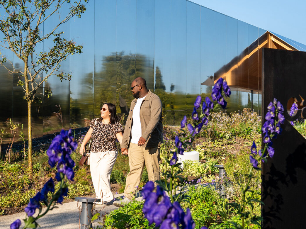 A couple walks hand-in-hand past a mirrored building and blooming purple flowers at Calder Gardens, with the garden's sign in the foreground.