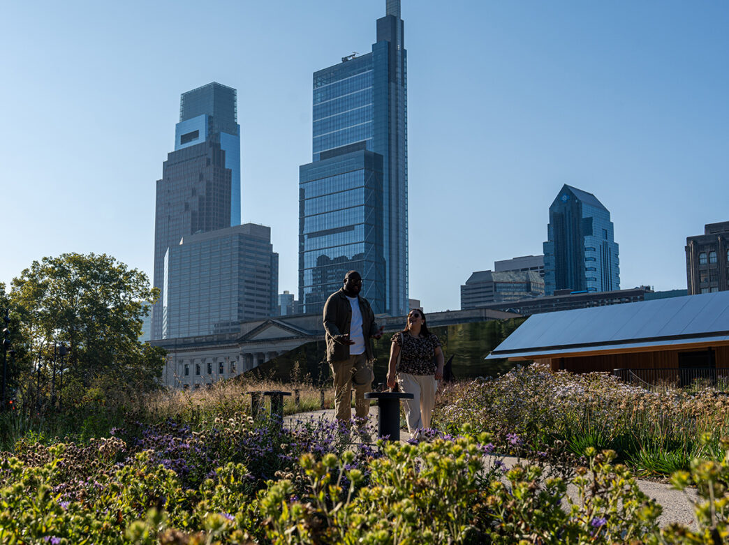 A could walks along a flower-lined path at Calder Gardens, with the city skyline in the background.