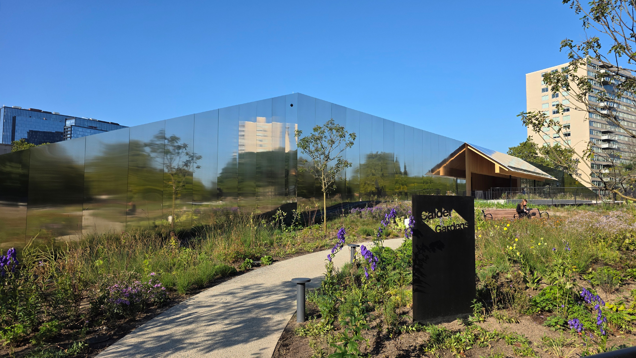 A mirrored building reflects the surrounding greenery, with a pathway lined by flowers leading to the entrance.