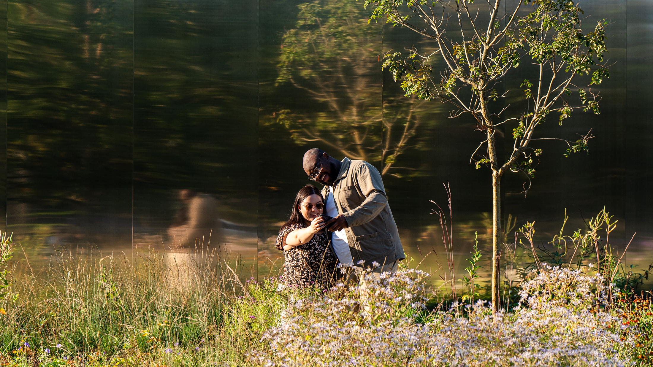 A couple stands close together among wildflowers in front of a mirror building while taking a selfie together.