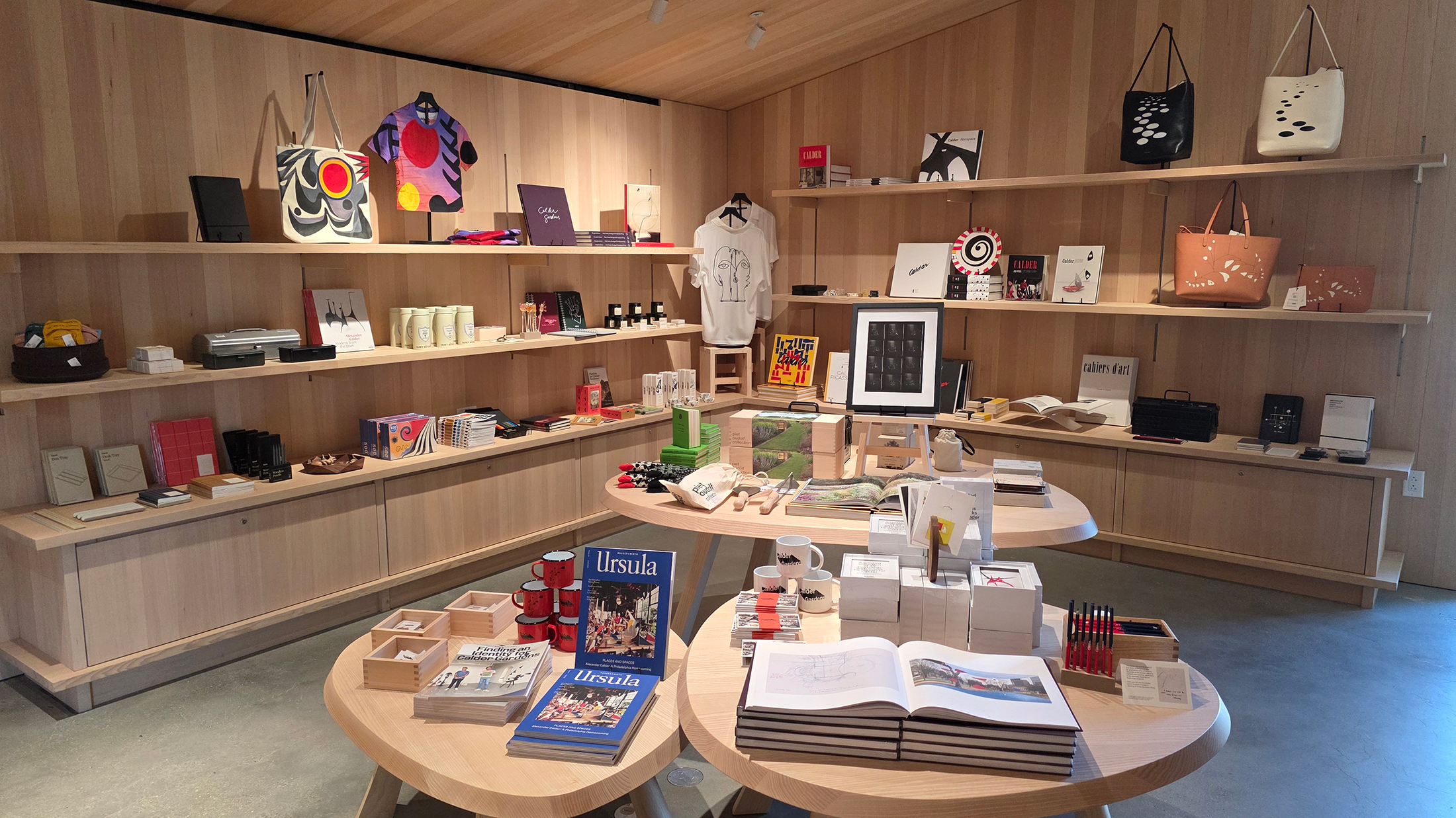 A gift shop with books, art-inspired tote bags, apparel and design objects neatly displayed on wooden shelves and tables.