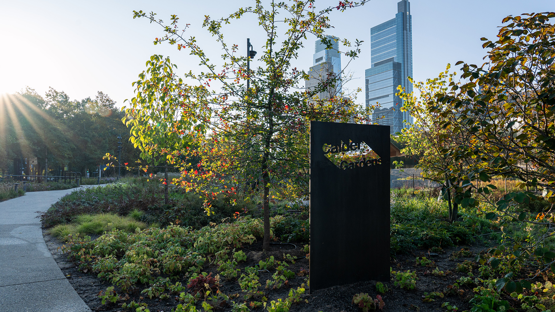A Calder Gardens sign sits among trees and paintings as sunlight filters through, with Philadelphia's skyline in the background.