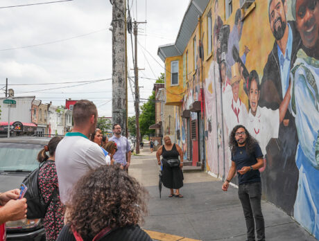 A guide speaks to a group of people gathered on a city sidewalk in front of a colorful mural depicting smiling figures, during a walking tour in the Centro de Oro neighbhorhood.