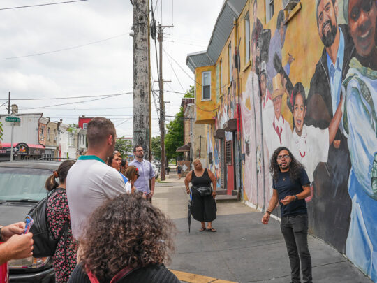 A guide speaks to a group of people gathered on a city sidewalk in front of a colorful mural depicting smiling figures, during a walking tour in the Centro de Oro neighbhorhood.