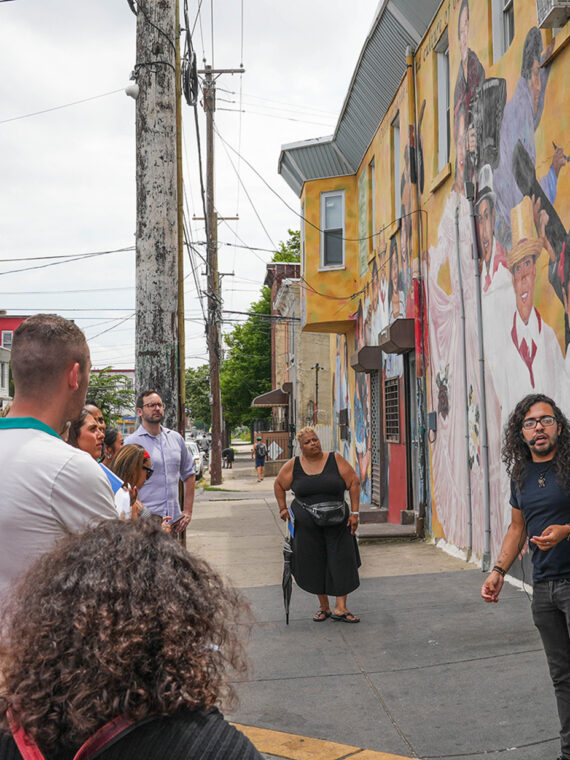 A guide speaks to a group of people gathered on a city sidewalk in front of a colorful mural depicting smiling figures, during a walking tour in the Centro de Oro neighbhorhood.