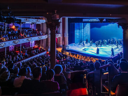 A wide shot inside the Academy of Music. The lights are turned down and the stage is lit up. Every seat in the theater is full.