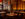 The elegant and warm dining room at Fork, featuring, artful wall murals resembling trees, an impressive bar display, amber chandeliers and tables set with wine glasses and candles for a cozy, upscale atmosphere.