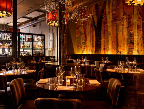 The elegant and warm dining room at Fork, featuring, artful wall murals resembling trees, an impressive bar display, amber chandeliers and tables set with wine glasses and candles for a cozy, upscale atmosphere.