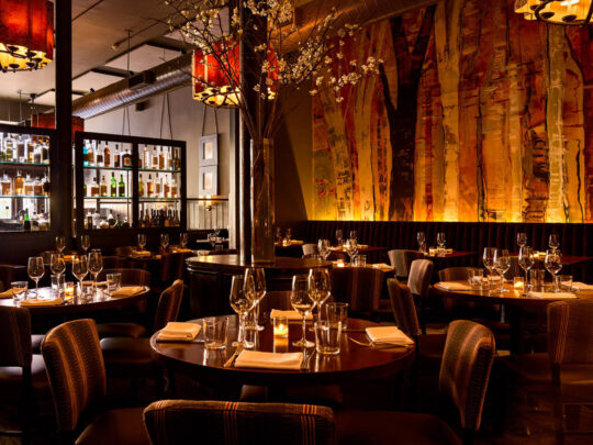 El elegante y cálido comedor de Fork, con murales artísticos que parecen árboles, una impresionante barra, lámparas de araña de color ámbar y mesas con copas de vino y velas para crear un ambiente acogedor y de lujo.