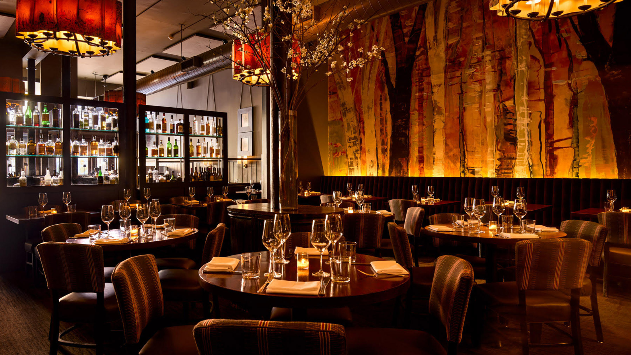 The elegant and warm dining room at Fork, featuring, artful wall murals resembling trees, an impressive bar display, amber chandeliers and tables set with wine glasses and candles for a cozy, upscale atmosphere.
