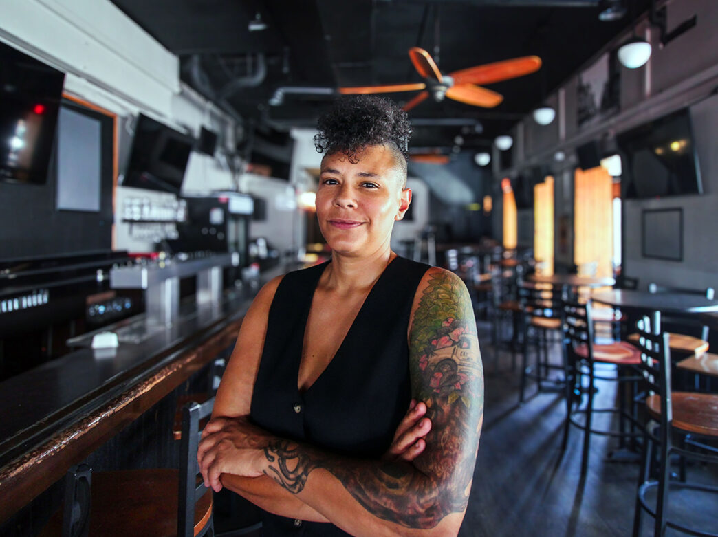 A smiling bar owner with tattooed arms stands confidently with folded arms inside a dimly lit bar with wooden high-top tables.