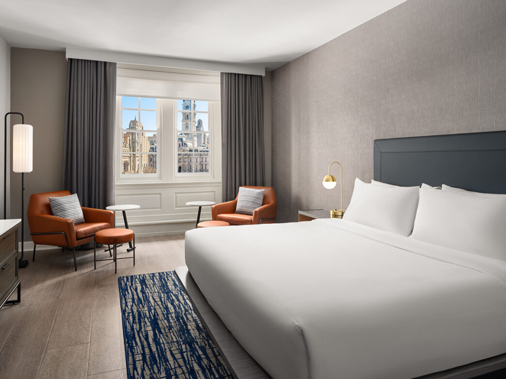 A hotel guestroom with a king bed with white linens, two orange accent chairs and a window overlooking Philadelphia's City Hall.