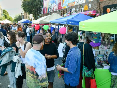 Crowds at a lively block party chat, browse vendors and enjoy the atmosphere. Colorful vender tents decorated with balloons line the car free street.