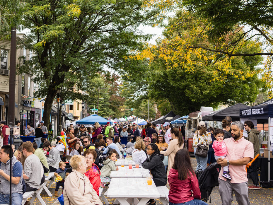 Crowds gather at long picnic tables and vendor tents during an outdoor street festival, with people eating, shopping and enjoying the lively atmosphere.