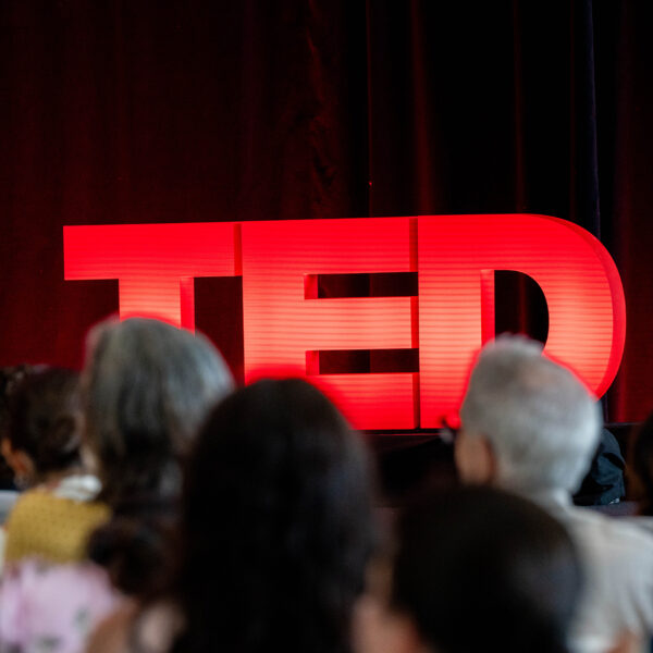 Audience members face a stage where a bright red TED sign glows against a dark red curtain backdrop.