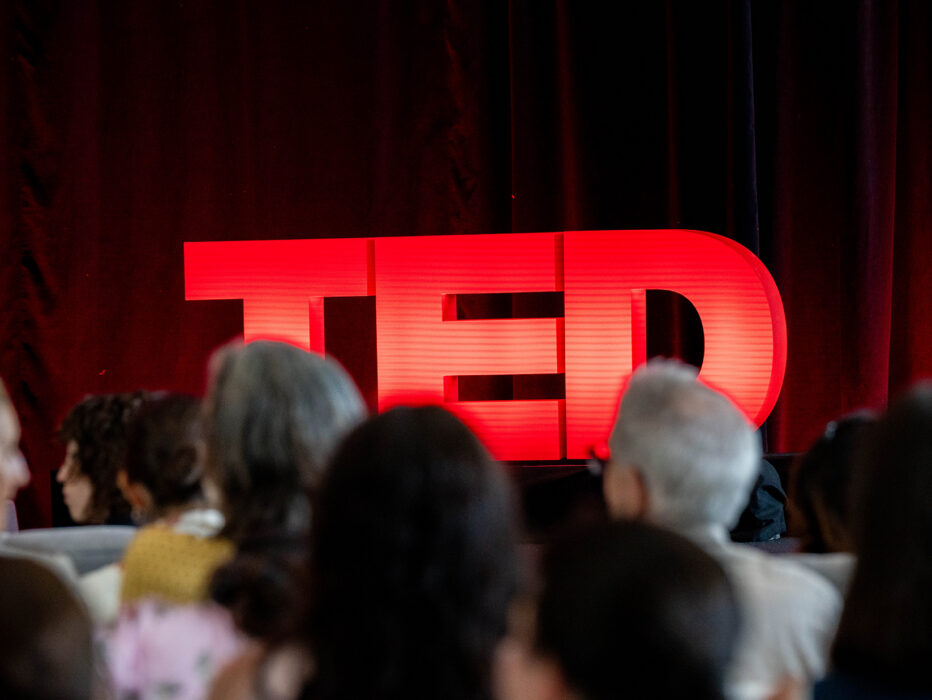 Les membres du public font face à une scène où un panneau TED rouge vif brille sur un fond de rideau rouge foncé.