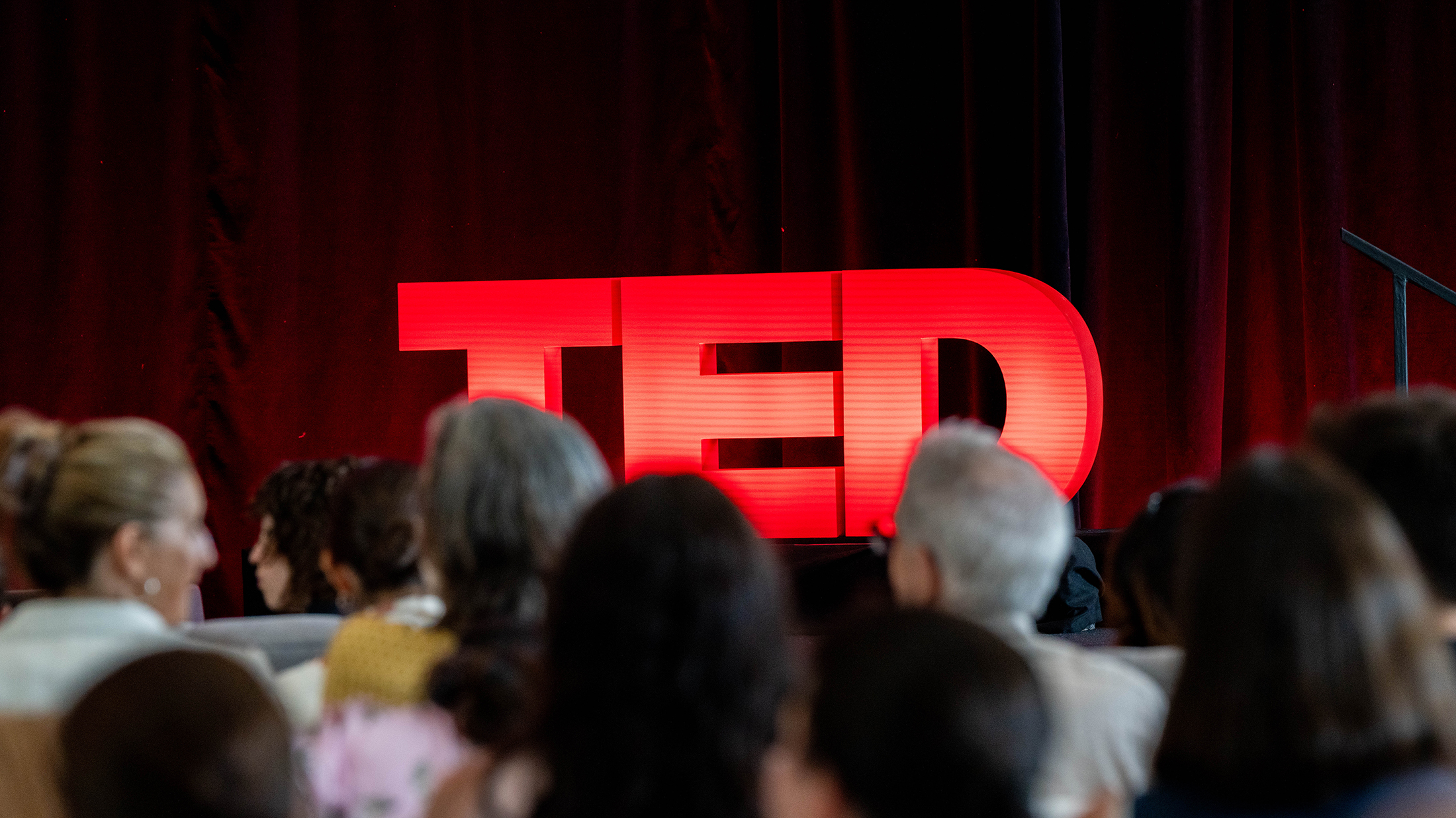 Audience members face a stage where a bright red TED sign glows against a dark red curtain backdrop.