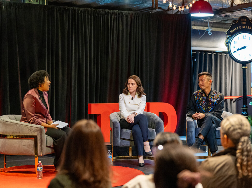 A moderator leads a conversation with two speakers on stage in front of a seated audience during a TED Democracy Fireside Chat inside Reading Terminal Market.