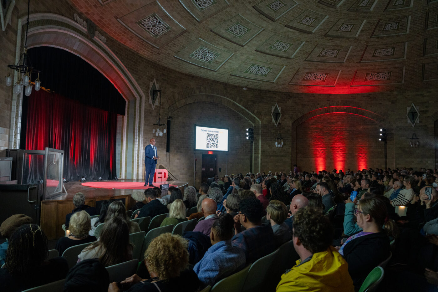A man stands on stage during a TED Talk as a large crowd looks on.
