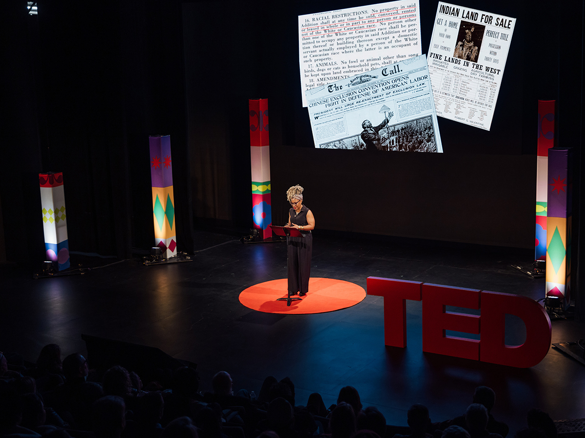 A speakers presents on stage with archival newspaper clippings projected on a screen behind them during a TED Democracy Speaker Series event.