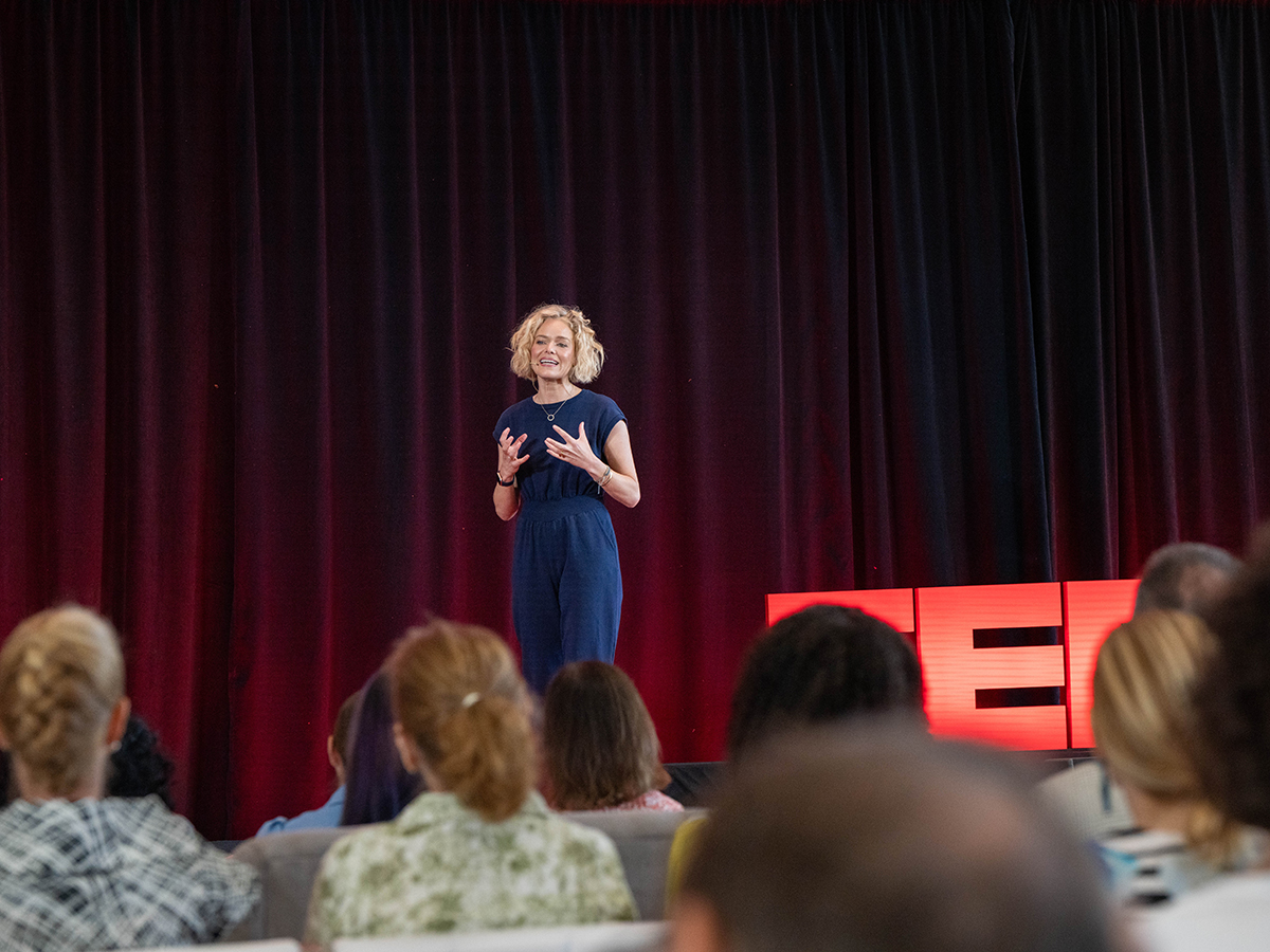 A woman in a blue jumpsuit speaks in front of a seated audience during a TED Democracy Fireside Chat event, standing near a red TED sign against a dark red curtain backdrop.