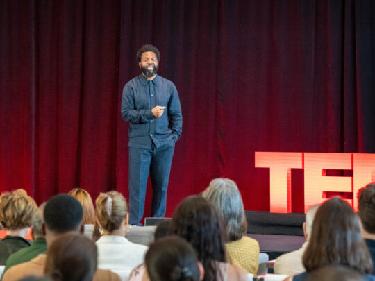 A man in a blue suit speaks in front of a seated audience during a TED Democracy Fireside Chat event, standing near a red TED sign against a dark red curtain backdrop.