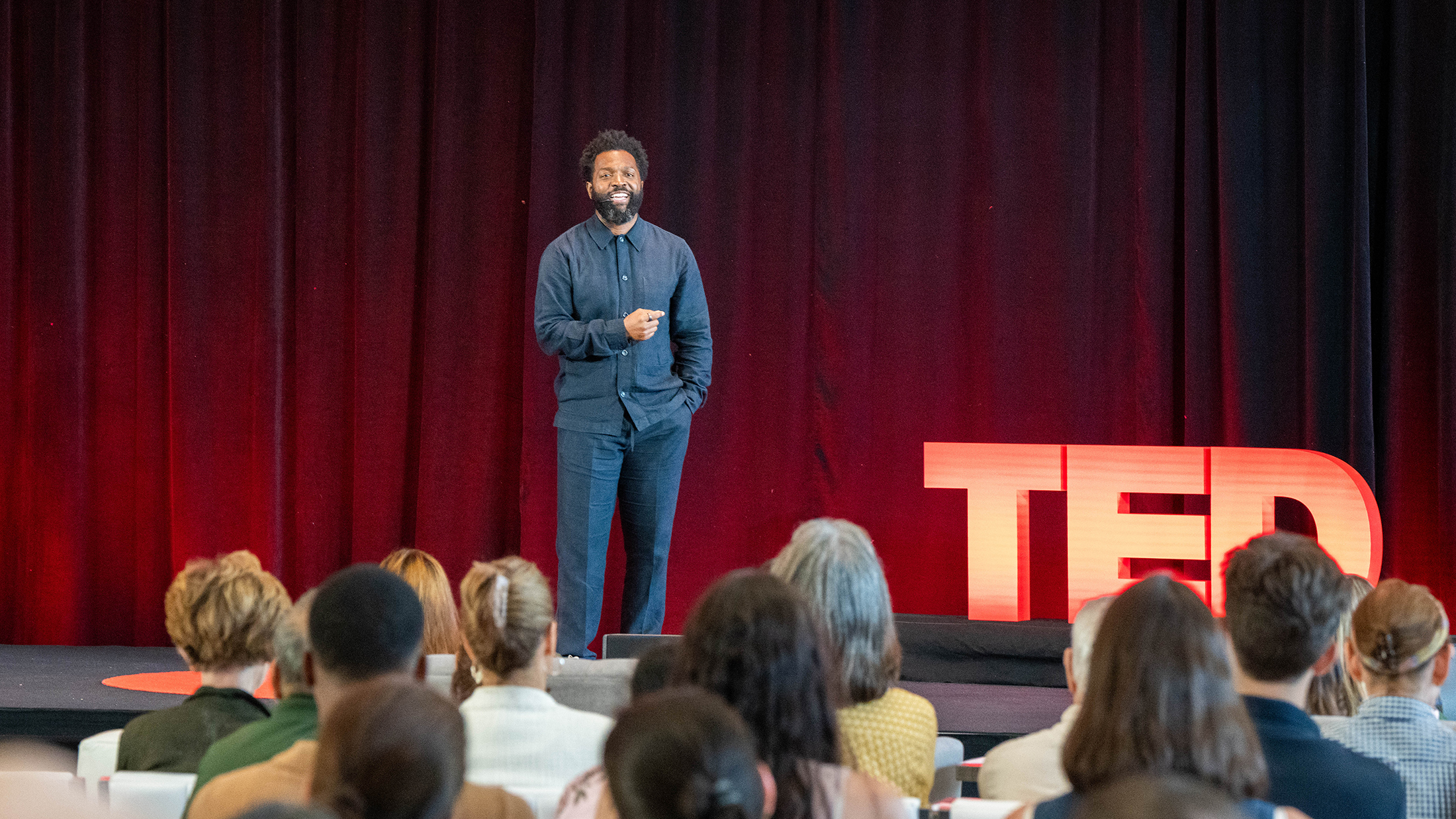 A man in a blue suit speaks in front of a seated audience during a TED Democracy Fireside Chat event, standing near a red TED sign against a dark red curtain backdrop.