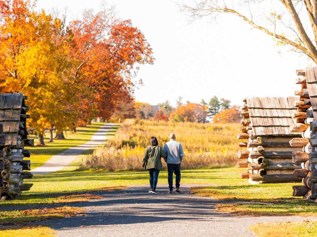 A couple strolls arm-in-arm down a pathway at Valley Forge National Historical Park, surrounded by vibrant fall trees and historic wooden huts.