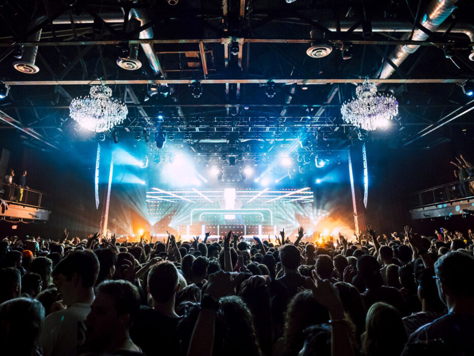 Una multitud con las manos en alto disfruta de un concierto lleno de energía bajo las brillantes luces del escenario y las lámparas de araña en The Fillmore.