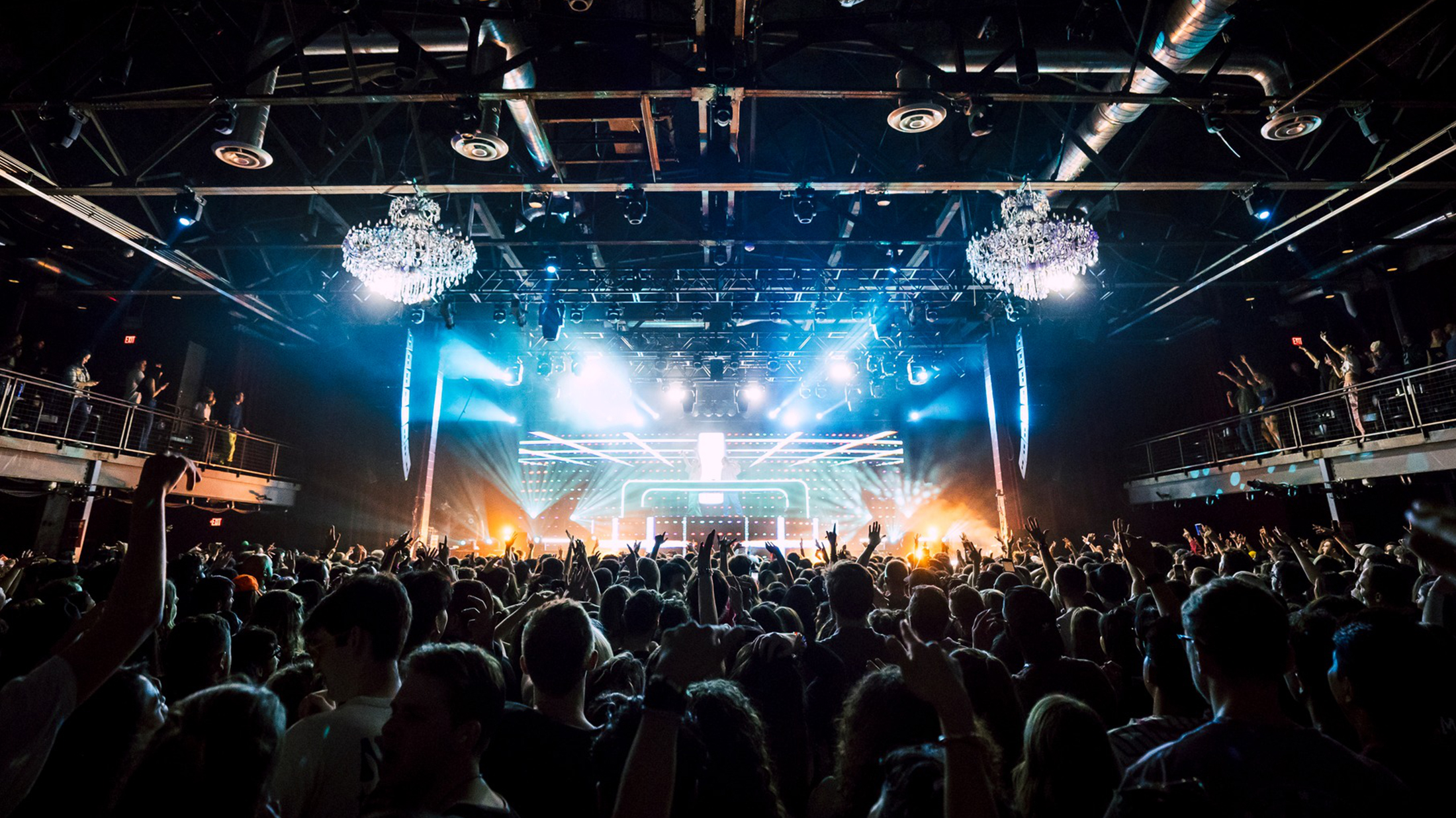 A packed crowd with hands raised overhead enjoy a high-energy concert under bright stage lights and chandeliers at The Fillmore.