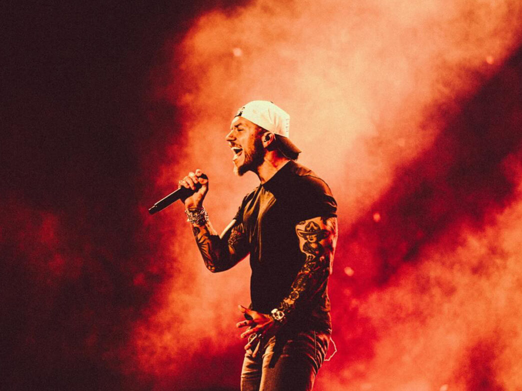 A man wearing a black t-shirt and backwards white baseball hat sings into a microphone with red and black stage smoke behind him.