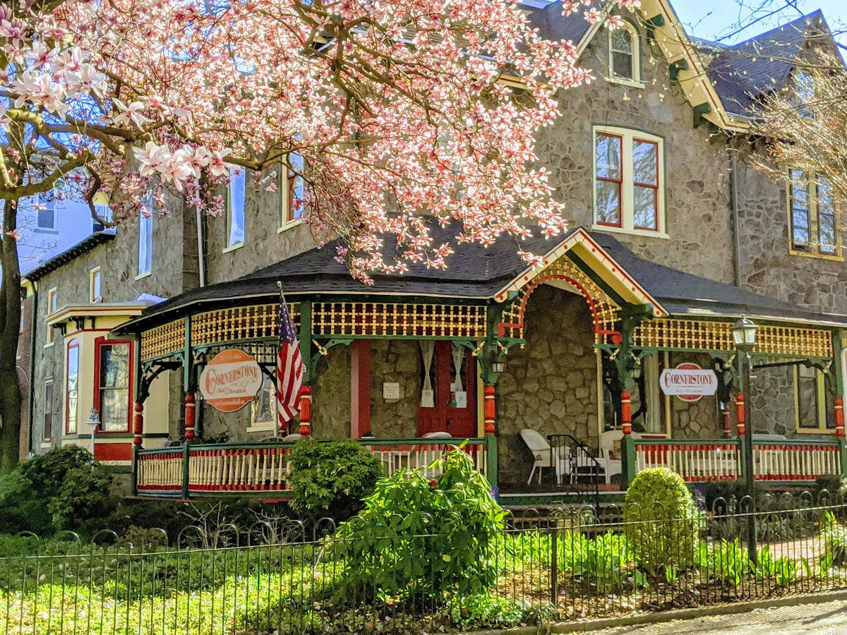A historic stone home with a wraparound porch, colorful trim and a blooming pink tree in spring.