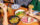 A group of diners sharing Asian-inspired dishes at a wooden table, with bao buns, French fries and roasted mushroom dumplings. Hands holding chopsticks reach to grab food from the plates of food.